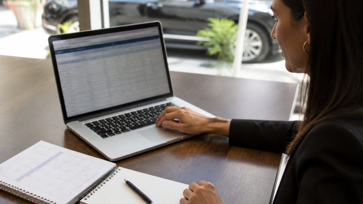 A travel manager at a desk reviewing a recurring ride schedule on a laptop, with a calendar and notepad showing weekly pickup times beside the keyboard