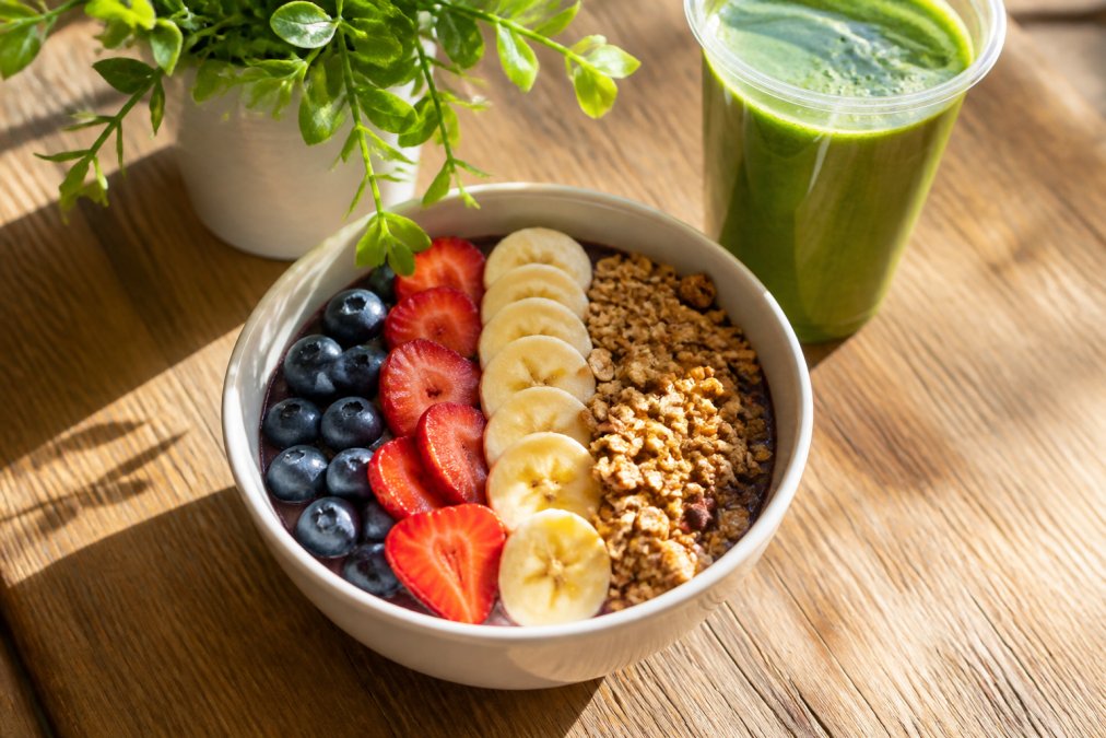 A bright overhead shot of a colorful nutrient-dense breakfast bowl with fresh berries, sliced banana, granola, and a green smoothie on a wooden table in natural morning light