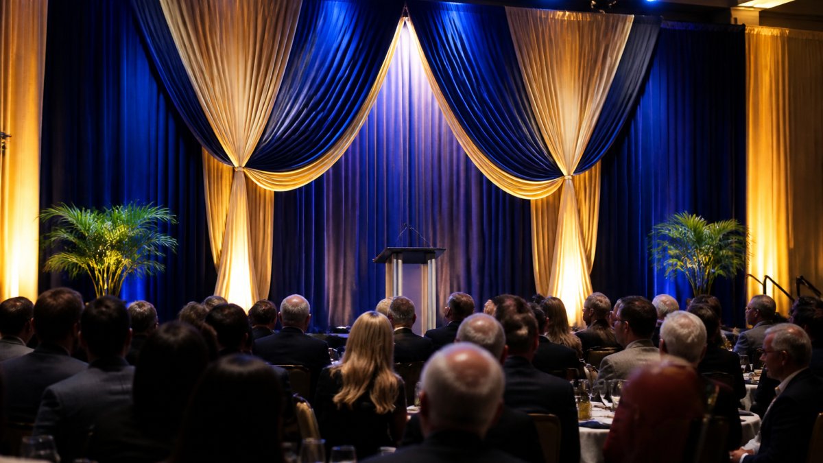 Professional corporate event stage with layered custom draping in deep blue and gold brand colors, large company logo prominently displayed on center backdrop panel, professional stage lighting illuminating the fabric, attendees seated in foreground