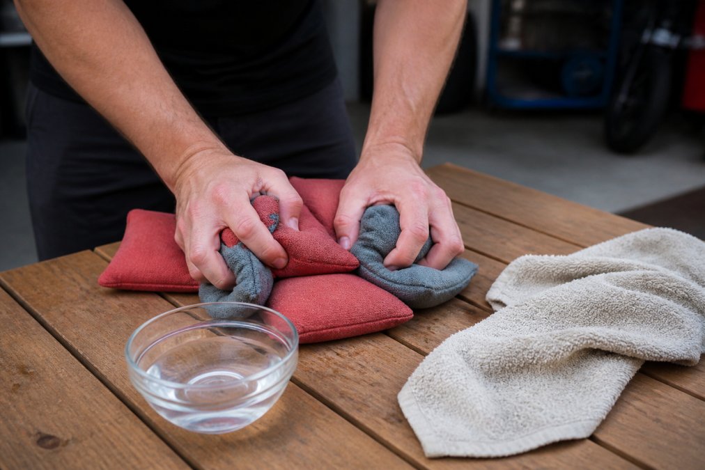 Close-up of hands kneading and massaging a set of cornhole bags on a wooden table, with a small bowl of distilled white vinegar and a towel nearby in a garage or backyard setting