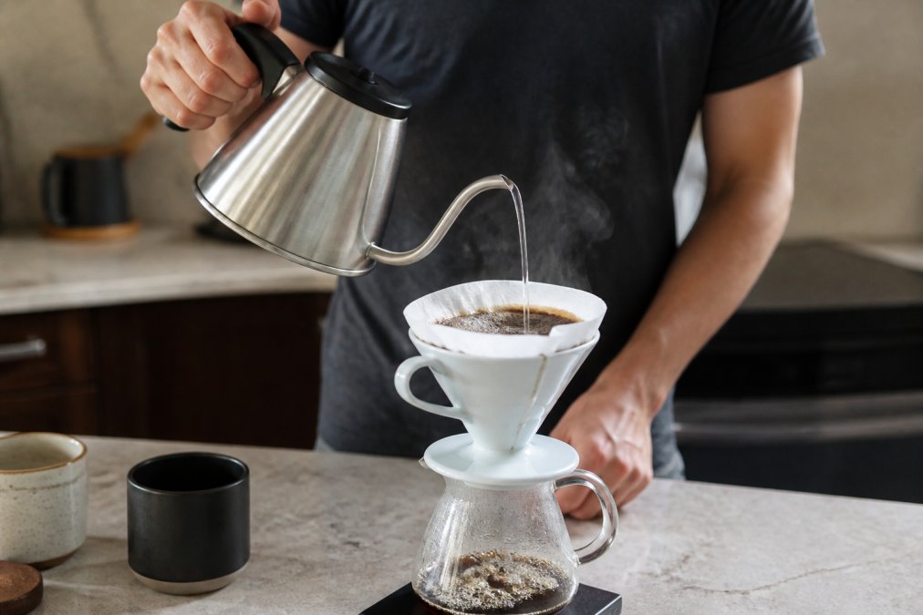 Hands pouring hot water from a gooseneck kettle into a pour-over dripper filled with medium-fine coffee grounds, showing controlled circular pouring motion and steam rising, natural kitchen lighting
