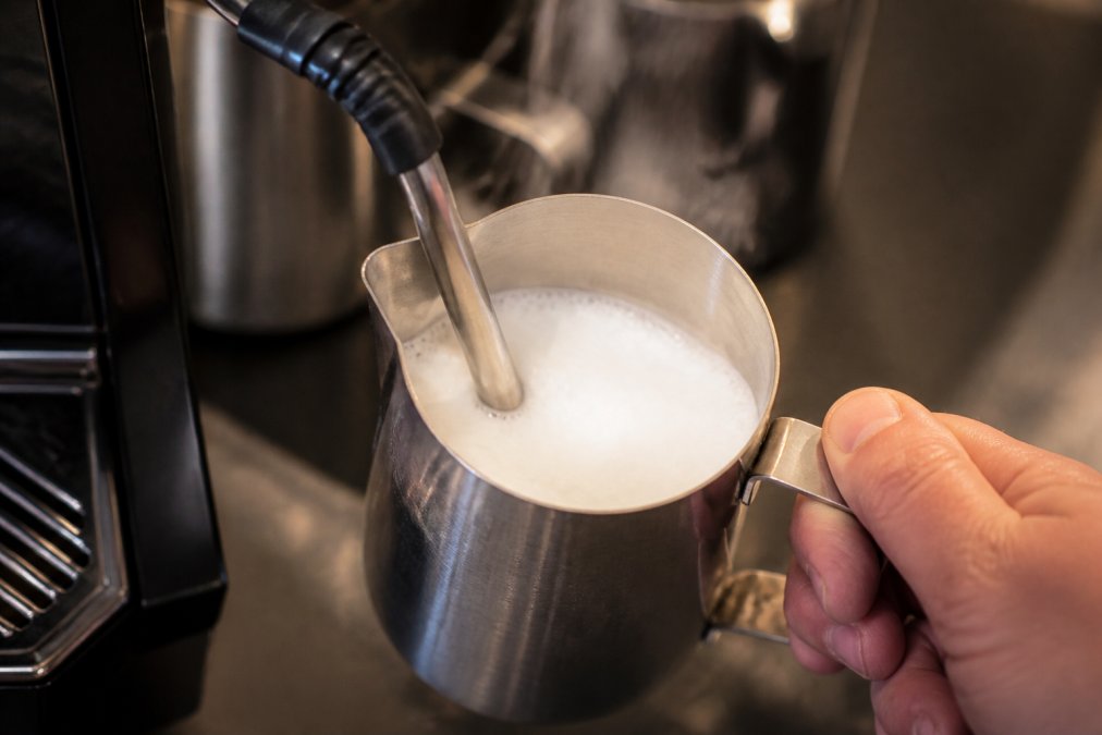 Close-up of a steaming wand positioned in a metal pitcher of milk, creating fine microfoam with visible texture on the milk surface, steam rising, professional espresso bar lighting