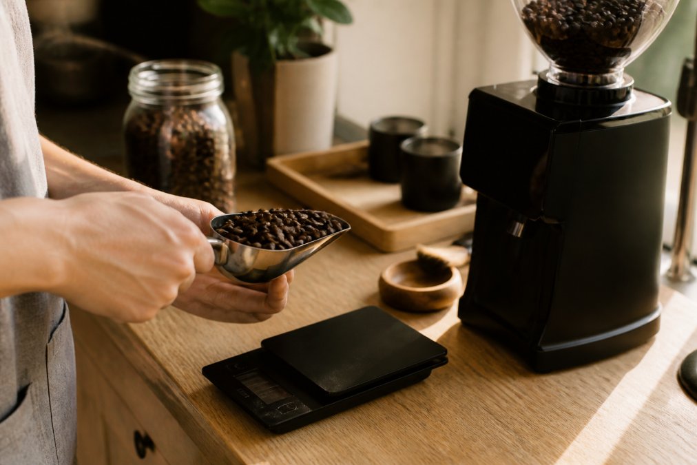 A person's hands holding a scoop of whole coffee beans above a digital scale next to a burr grinder on a wooden kitchen counter, warm morning light