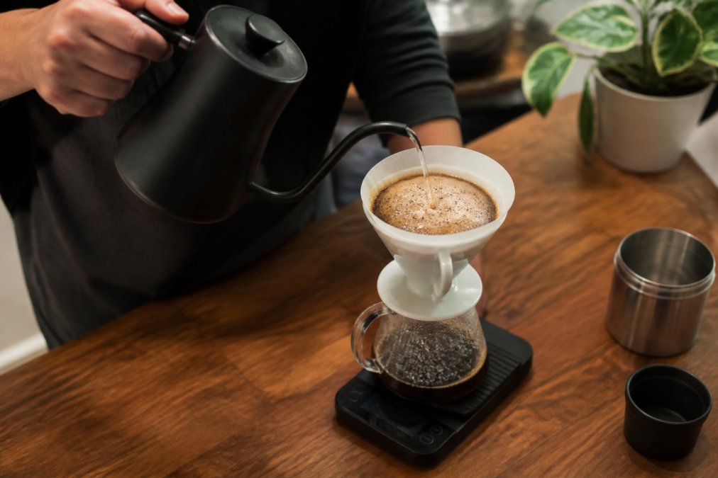 Close-up of hands carefully pouring hot water from a matte black gooseneck kettle over a pour over dripper with a white paper filter, coffee grounds blooming and rising in a dome shape, set on a wooden countertop with a digital scale visible underneath displaying 200 grams