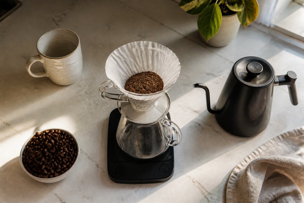 Overhead shot of a kitchen counter with a digital scale, a pour-over dripper, a gooseneck kettle, and a small bowl of whole coffee beans, morning light streaming in