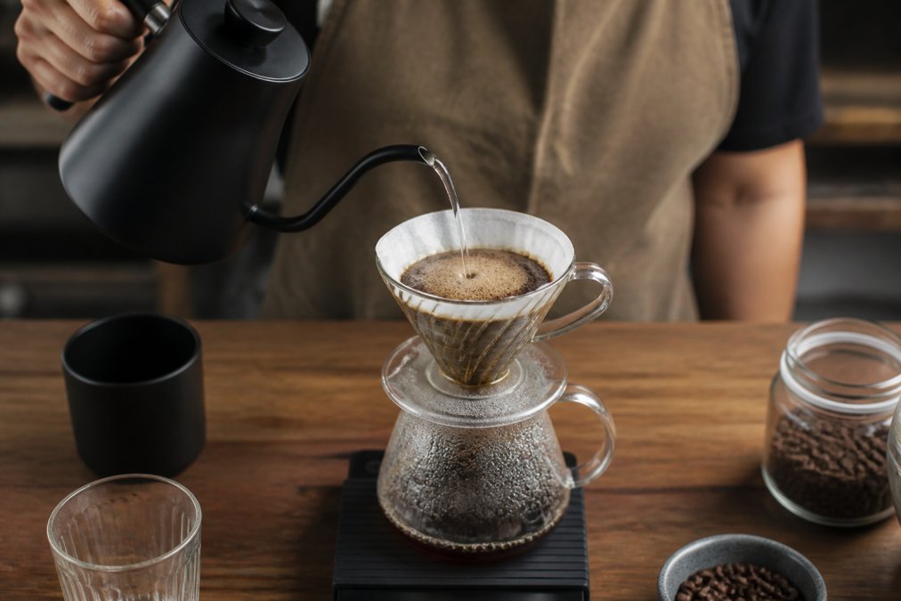Close-up of a hand pouring hot water from a gooseneck kettle in a slow, circular motion over a glass pour-over brewer with fresh coffee grounds blooming