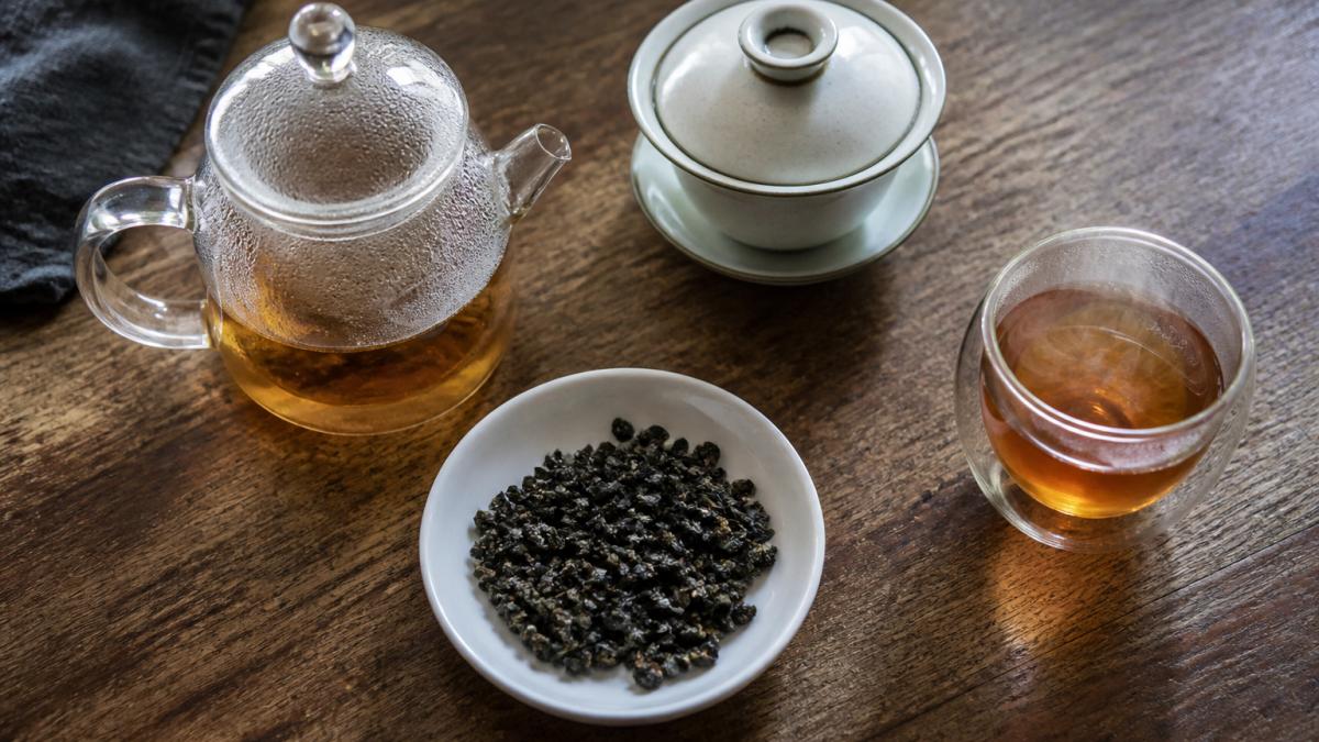 Glass teapot, a gaiwan, and loose tea leaves on a wooden counter with a steaming cup of amber oolong tea.