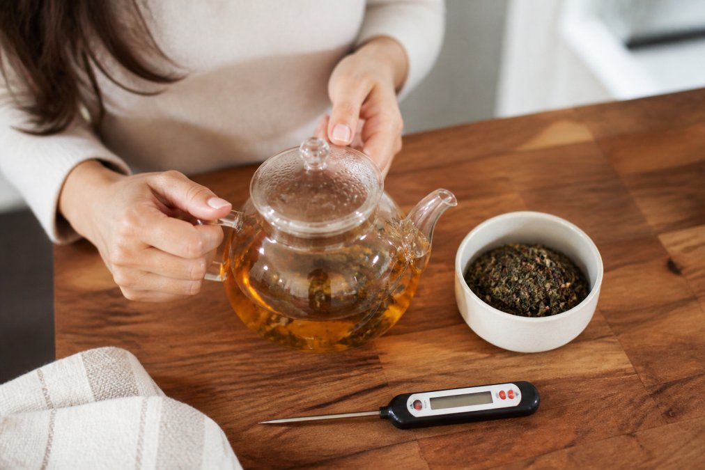 Glass teapot with amber tea and loose leaves beside a thermometer to help you brew loose leaf tea accurately.