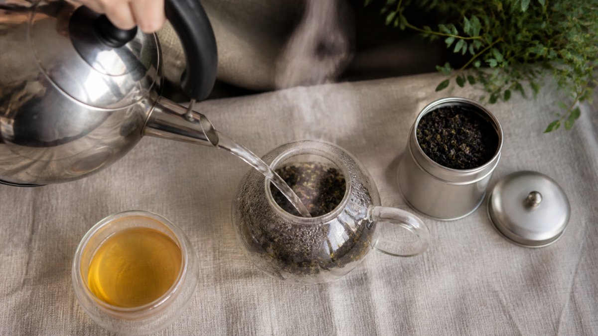 Overhead shot of hands pouring hot water from a kettle into a glass teapot filled with loose leaf tea, steam rising, a small metal caddy and a filled teacup on a linen-covered table beside it