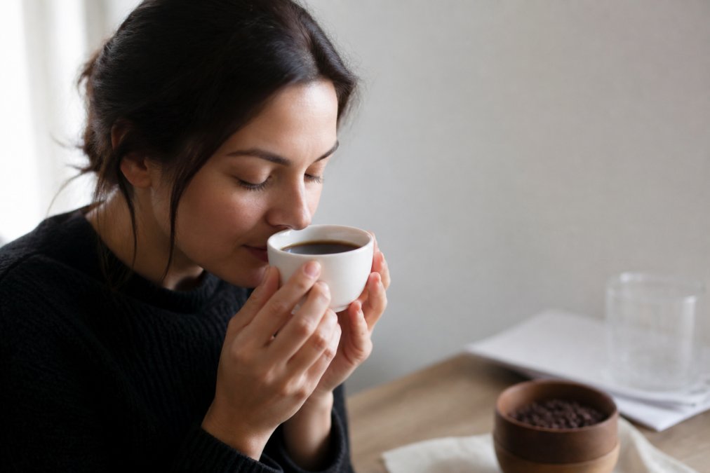 A person holding a white ceramic cup of black coffee close to their nose with eyes closed, savoring the aroma, soft natural light, minimalist background suggesting a quiet morning ritual