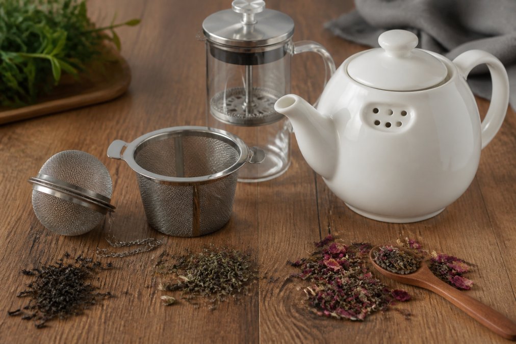 Flat lay of several tea infuser types arranged on a warm wooden surface, a stainless steel mesh ball infuser, a wide basket strainer, a glass French press, and a white ceramic teapot with a built-in strainer, surrounded by scattered loose leaf tea leaves in natural daylight