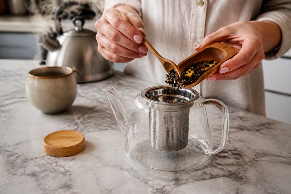 Close-up of a person's hands carefully spooning loose leaf tea from a small wooden scoop into a glass teapot's basket infuser, with a steaming kettle and a simple ceramic mug visible in the soft-focus background on a light marble kitchen counter