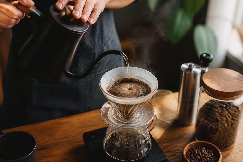Close-up of hands holding a gooseneck kettle, pouring hot water in a slow spiral over a pour-over dripper with a white filter and dark coffee grounds, steam rising gently from the surface, on a warm wooden countertop in soft morning light