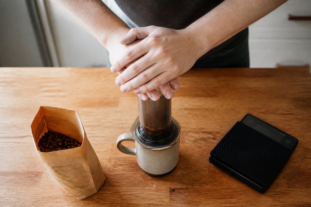 Overhead shot of hands pressing an AeroPress over a ceramic mug on a wooden kitchen counter, with a bag of coffee beans and a digital scale nearby