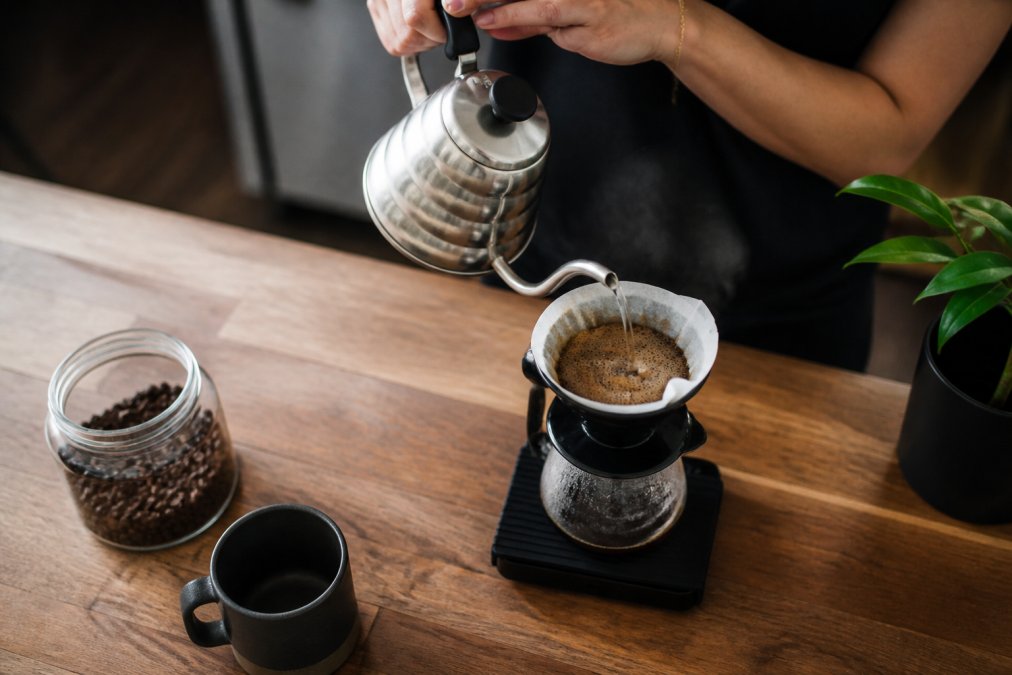 Hands pouring hot water from a gooseneck kettle to brew specialty coffee using a pour-over dripper on a scale.