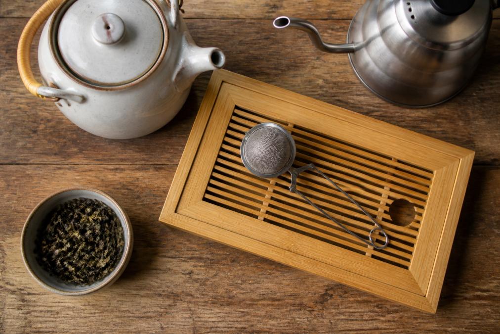 Flat lay of loose leaf tea brewing tools on a wooden wooden table: a ceramic teapot, bamboo tea tray, mesh infuser, gooseneck kettle, and small bowl of loose leaf tea leaves