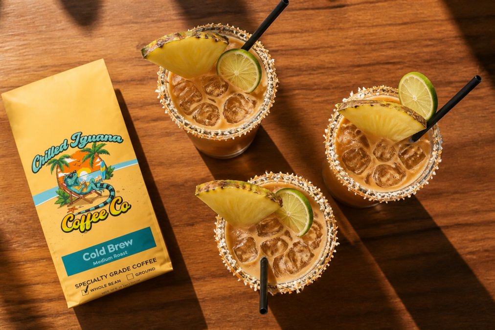 Overhead view of three colorful tropical coffee drinks in tall glasses with fresh pineapple slices, toasted coconut rim, and lime wheels as garnishes on a bright wooden table in afternoon sunlight