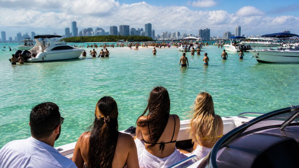 A group of friends on a private speedboat approaching a busy sandbar with clear turquoise water, people wading and relaxing, Miami skyline visible in the distance