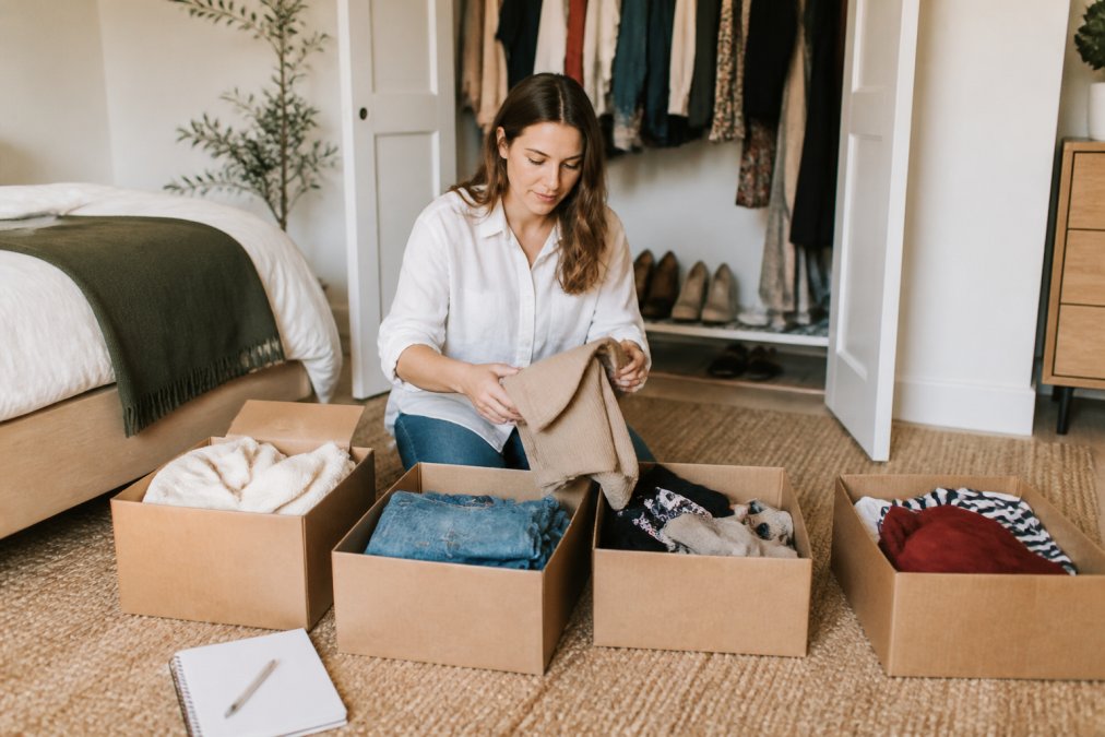 A woman in her thirties sitting on the floor of a bright bedroom, sorting clothes into four labeled boxes, with a closet visible behind her and a notepad nearby