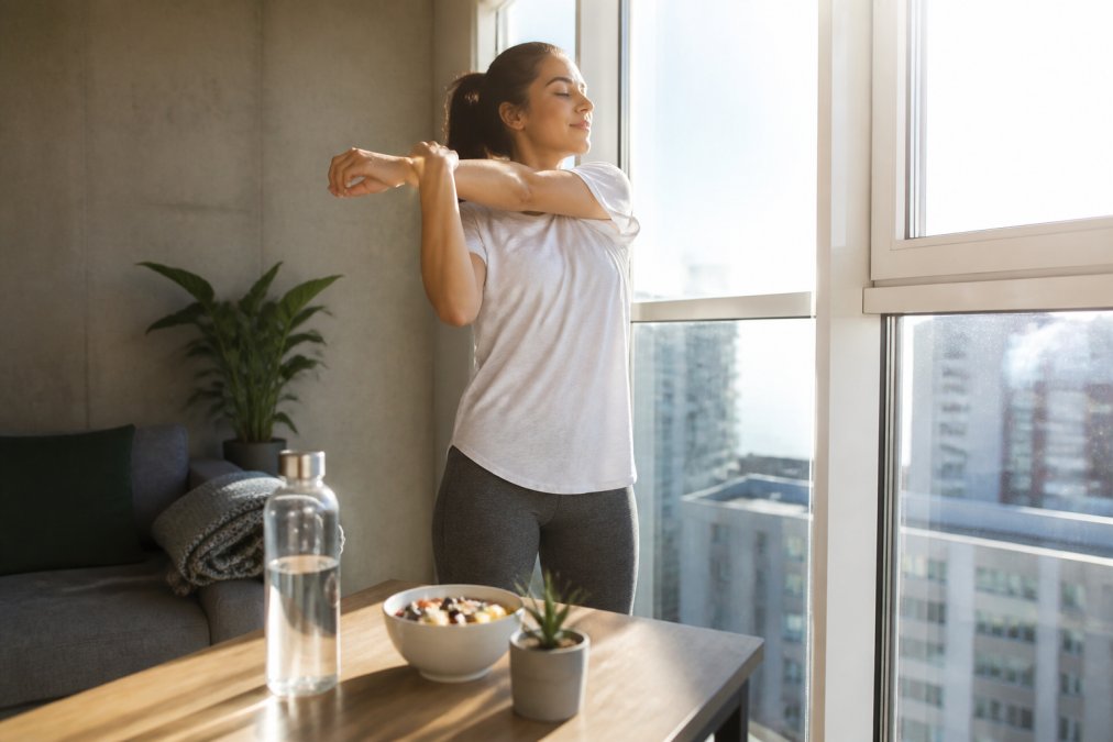 Person in morning sunlight doing gentle stretching near a window with a water bottle and healthy breakfast visible on a nearby table
