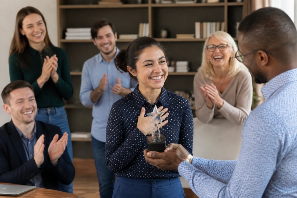 A diverse team visible on a video call, with one employee smiling while receiving recognition on screen, showing virtual team celebration with colleagues clapping or waving in support