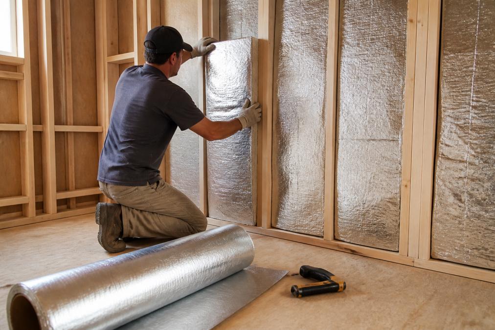 A person installing foil-faced insulation batts between wooden wall studs inside a partially framed sauna room, with a roll of vapor barrier and a staple gun nearby on the subfloor