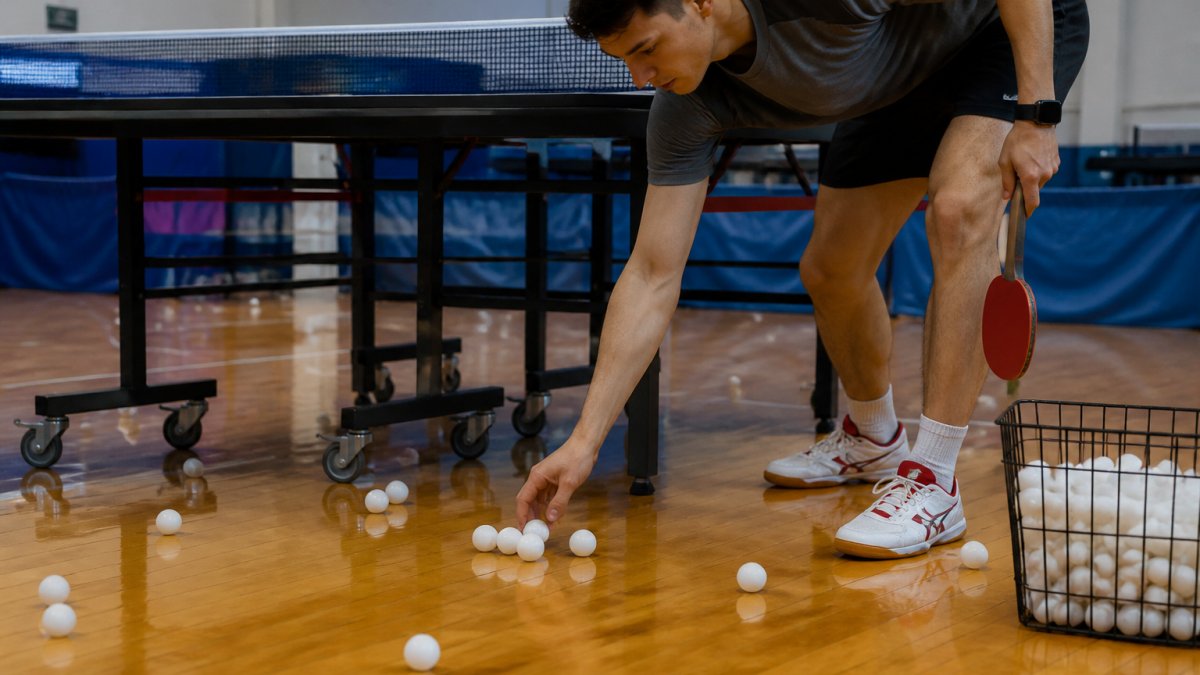 Player demonstrating efficient ball collection technique using a handheld scoop tool to gather multiple ping pong balls from the floor near a table tennis table, with natural gym lighting