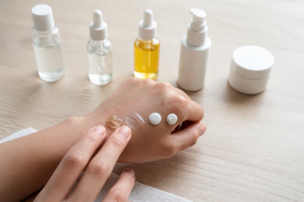 Hands demonstrating skincare product application on the back of the hand, showing bottles and serums arranged in order from lightest to heaviest consistency