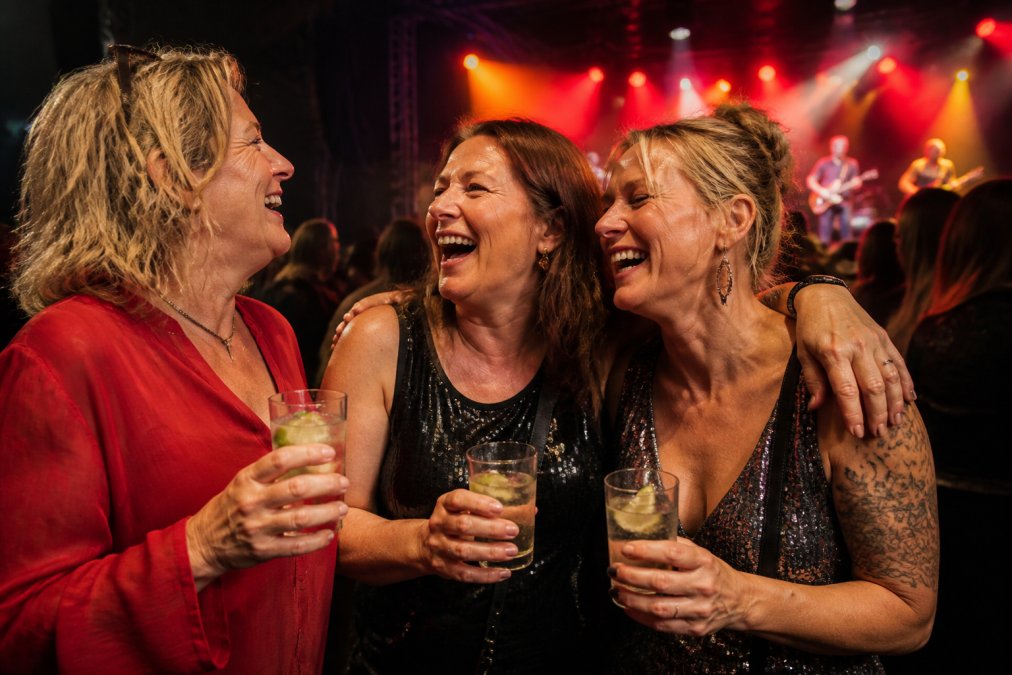 A group of women in their forties laughing and dancing together at a live music gig, holding sparkling water and lime in glasses, colourful lights behind them, dressed in bold going-out clothes
