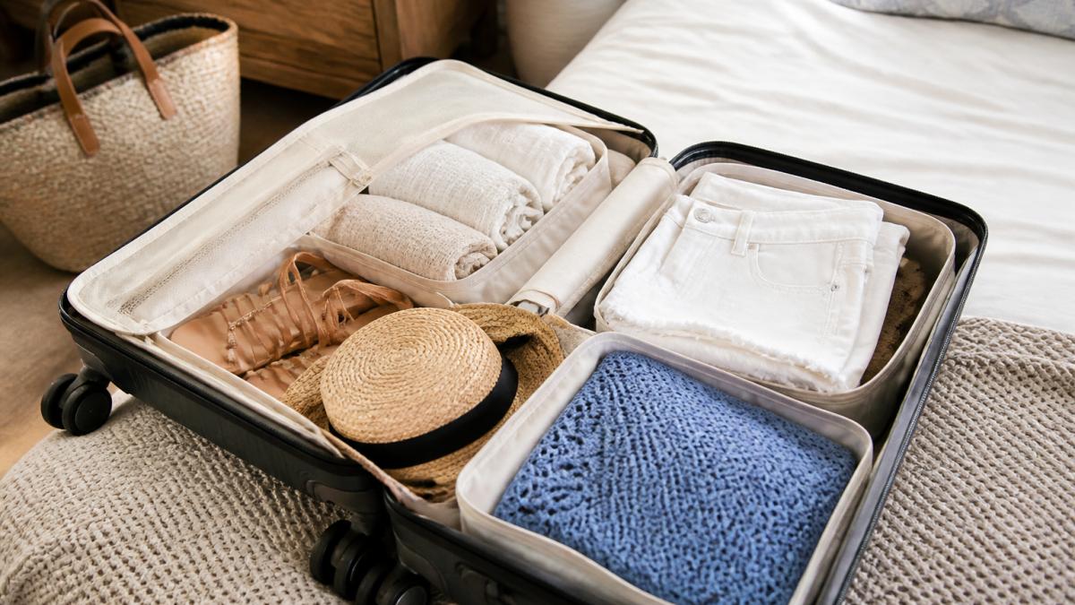 Open carry-on suitcase on a bed with rolled gauze dresses, folded white shorts, a blue open weave sweater, sandals, and a straw hat arranged in packing cubes