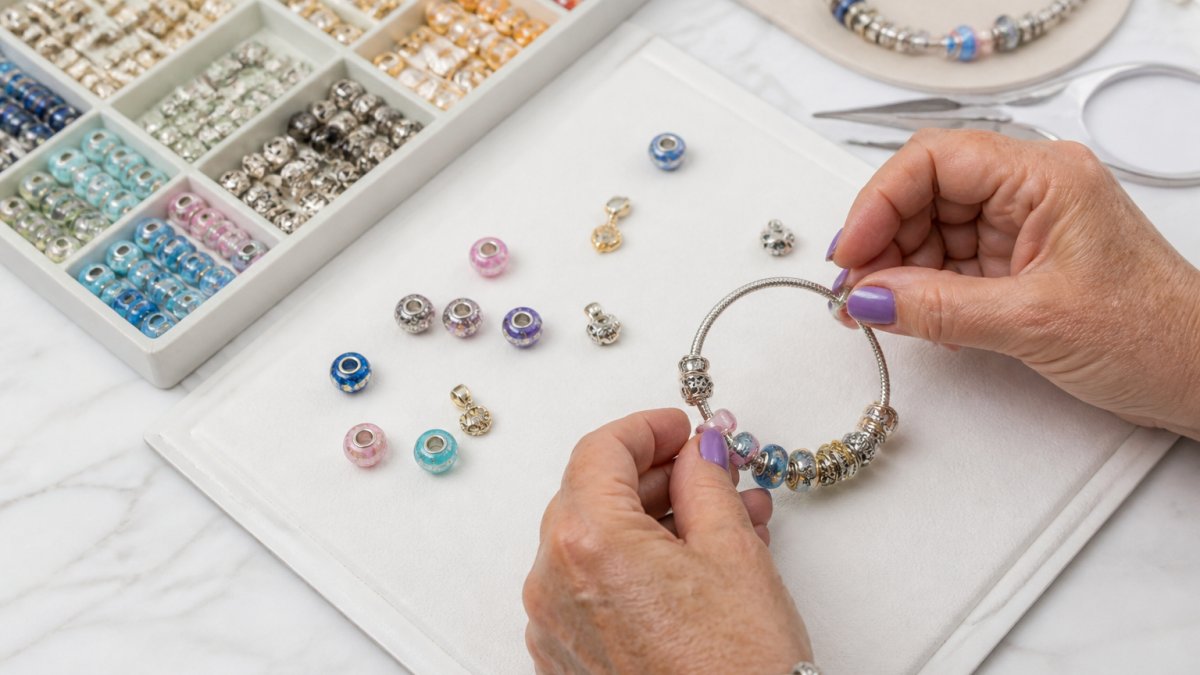 Close-up of hands arranging colorful glass and crystal beads on a beaded bracelet base, with organized materials and charms laid out on a white work mat