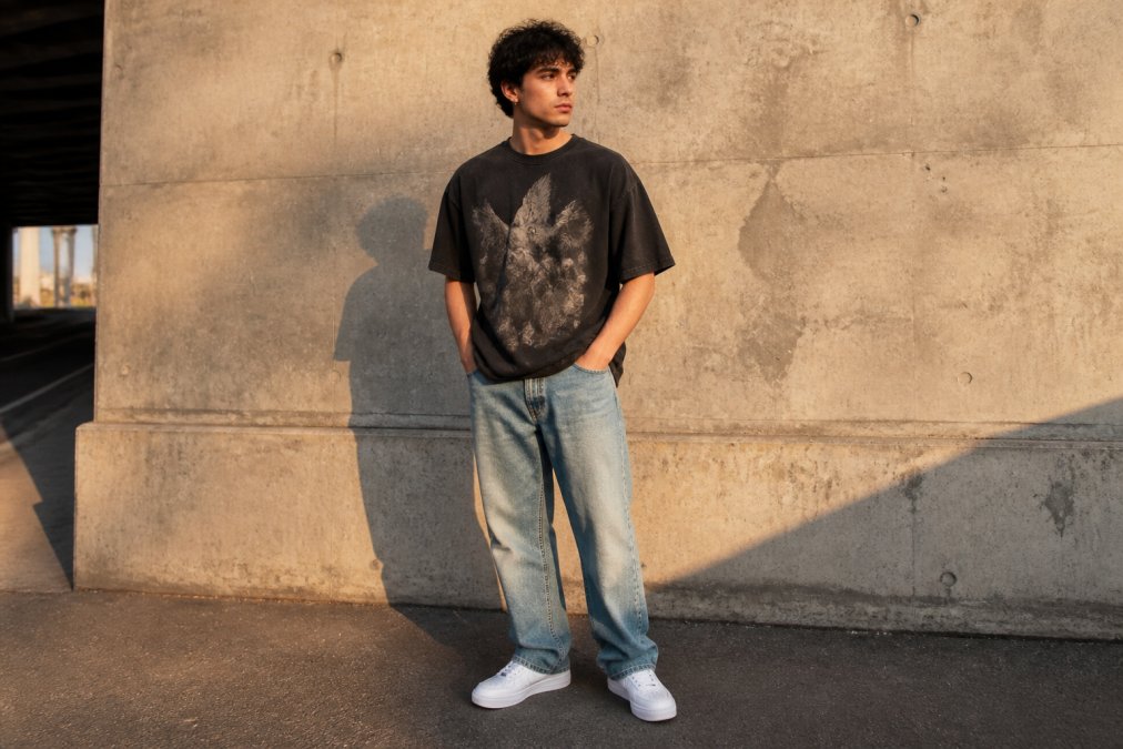 A young man wearing an oversized graphic t-shirt with straight-leg denim and clean white sneakers, standing against a concrete wall in an urban setting during golden hour