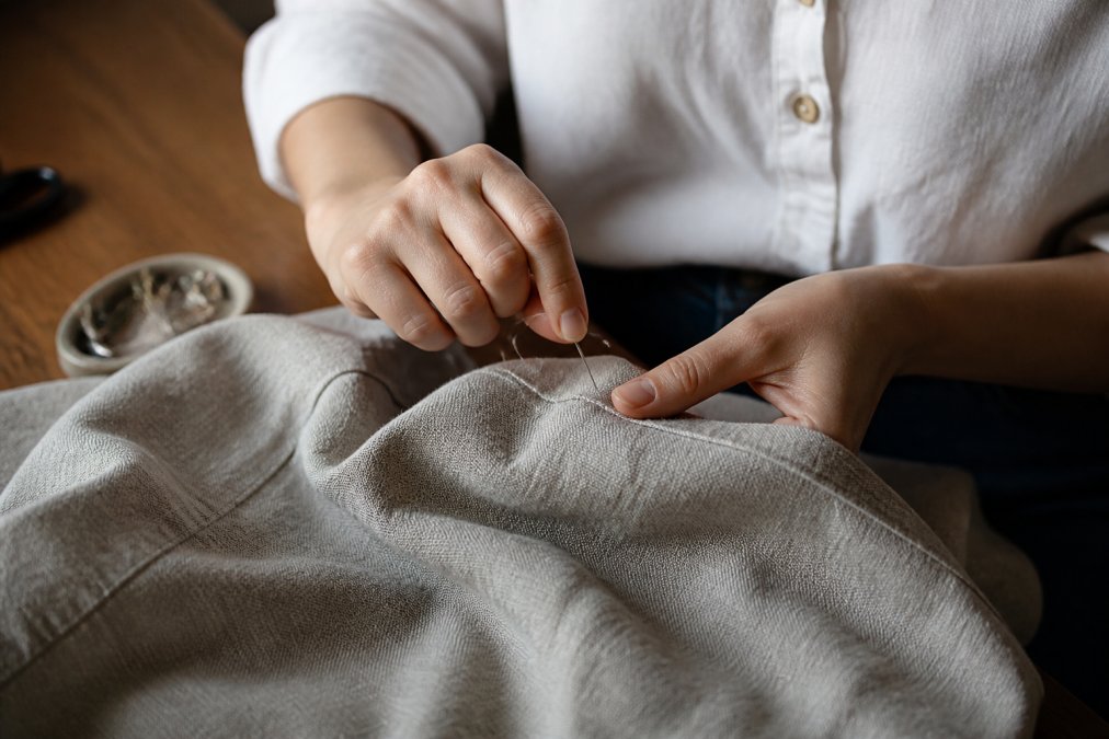 Close-up of hands carefully mending a natural fabric garment with needle and thread, showing detailed stitching work on linen material in warm indoor lighting