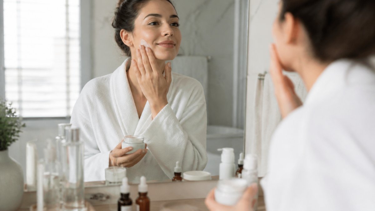 Woman applying skincare products in sequence on her face, showing hands with serums and moisturizers in a bright, clean bathroom