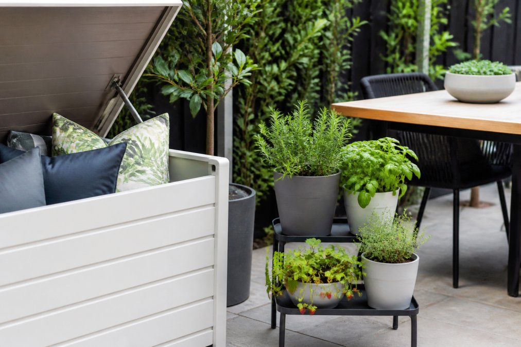 Close-up of a small entertaining area showing a weatherproof storage bench with cushions stacked neatly, potted herbs and edible plants arranged on tiered stands beside it, and a compact outdoor table in the background, photographed in natural daylight