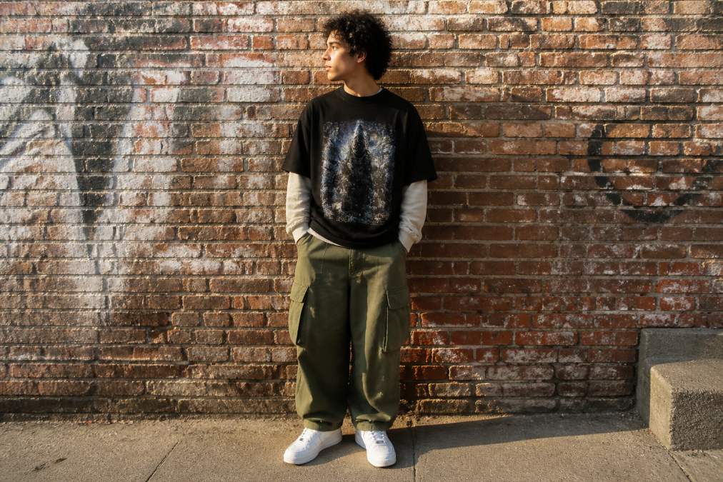 A young adult wearing an oversized vintage graphic tee layered over a long-sleeve shirt, paired with baggy cargo pants and clean sneakers, standing against a brick wall in warm afternoon light