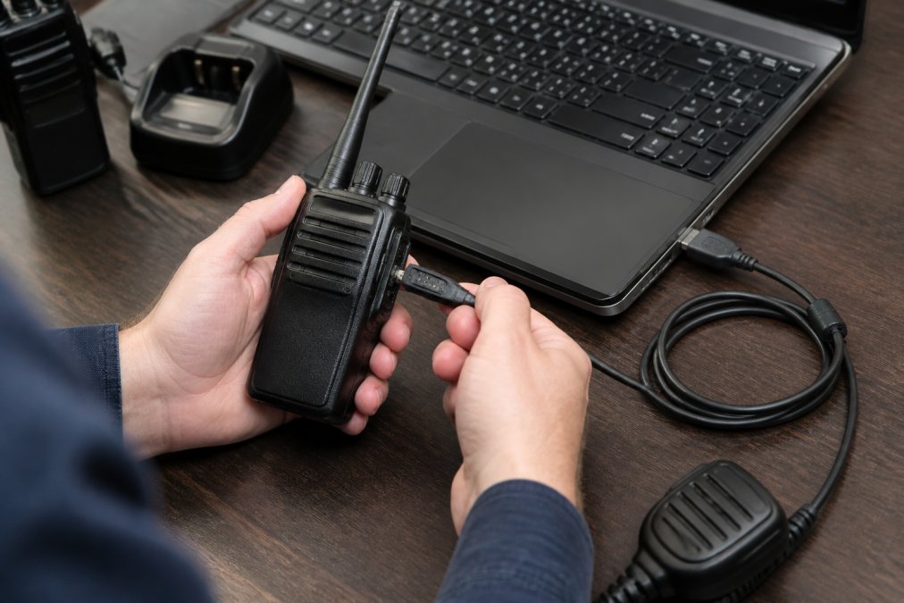Close-up of hands connecting a programming cable to a handheld transceiver next to a laptop displaying radio programming software interface