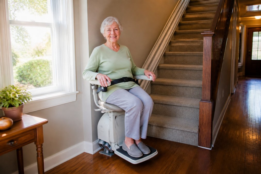 Older adult securely seated in a stairlift at the bottom of residential stairs, clearly showing the black seat belt fastened across the lap, padded armrests with control buttons, and the footrest extended, with natural afternoon light from a window