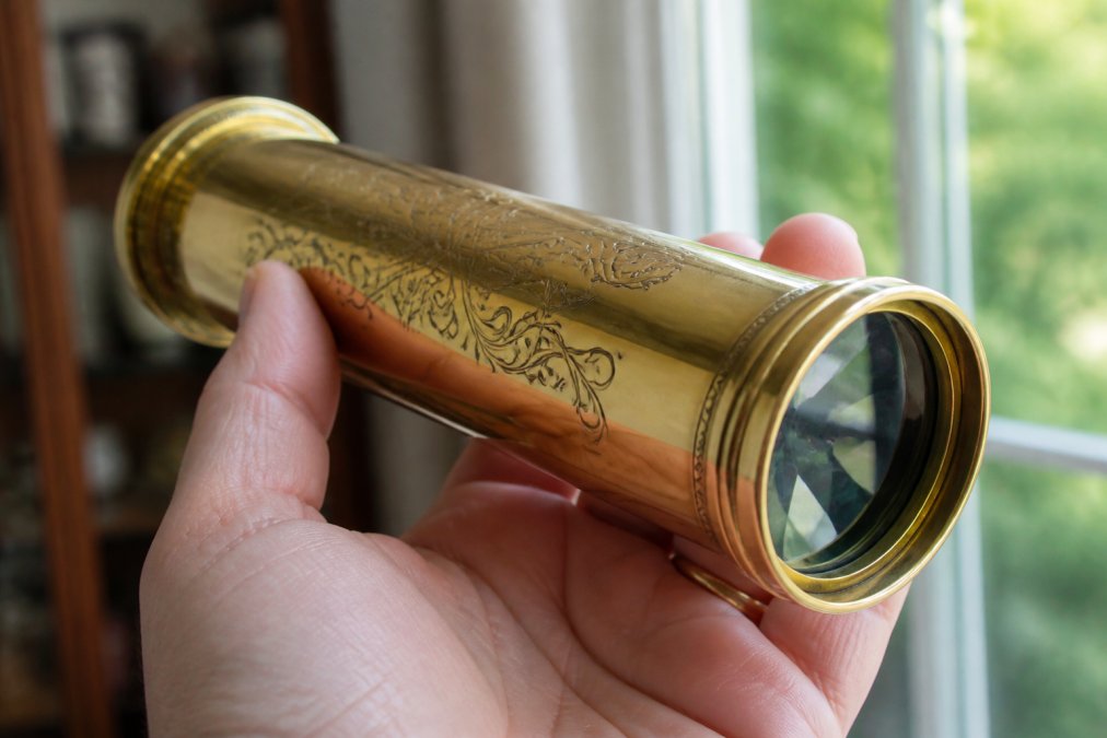 Close-up of a hand holding a polished brass kaleidoscope, showing the intricate acid-etched details and the reflective glass end, with soft window light illuminating the scene