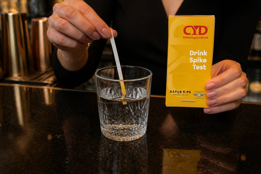 Close-up of hands holding a drink testing strip partially immersed in a clear beverage in a glass at a bar counter, with the strip beginning to show colour change against the liquid