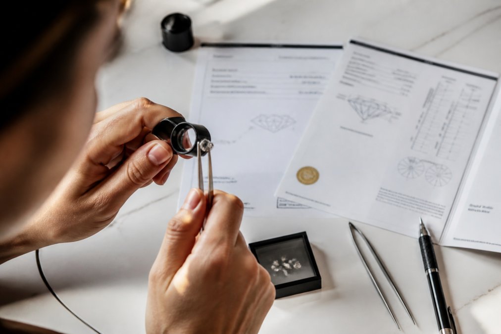 Close-up of hands examining a diamond with a jeweler's loupe, with a GIA certification document and diamond grading report visible on a white surface in natural daylight