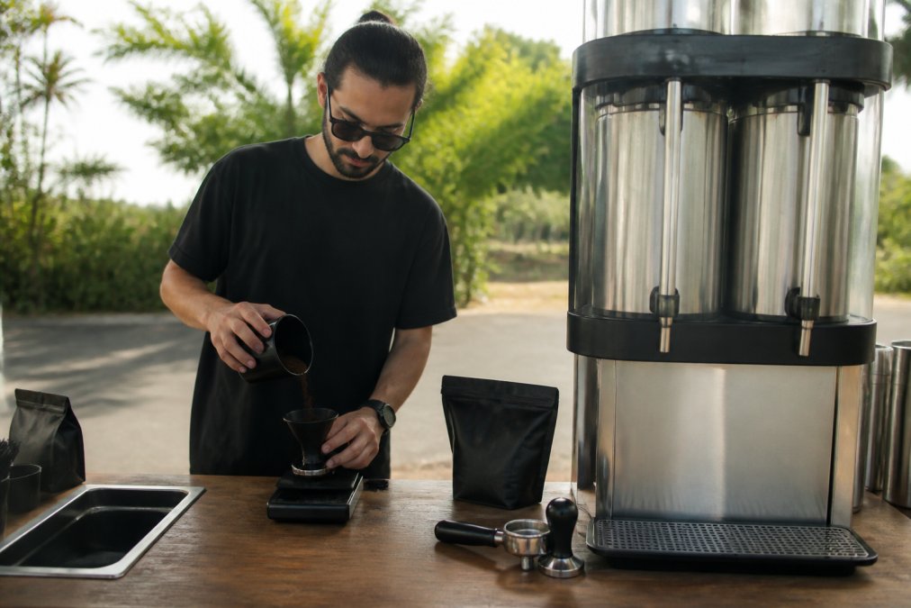 A barista measuring dark coffee grounds on a digital scale next to a large commercial brewer at an outdoor event setup, morning light