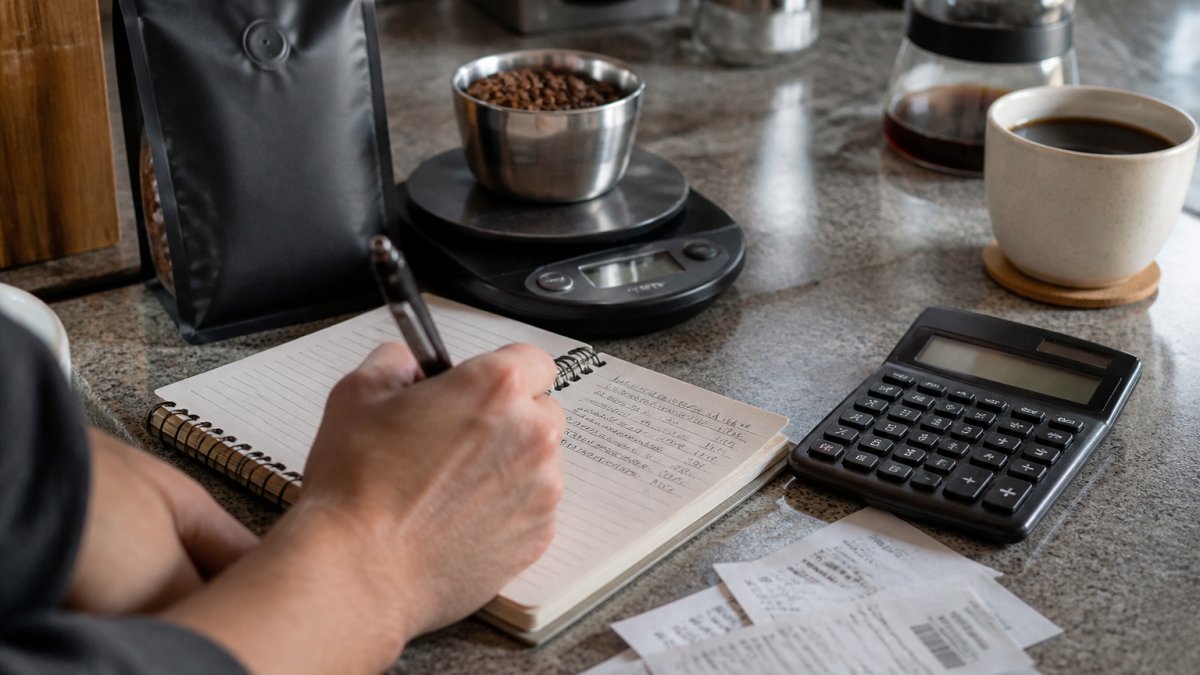 Hands writing measurements in a notebook to learn how to calculate cost per cup of home coffee on a kitchen counter