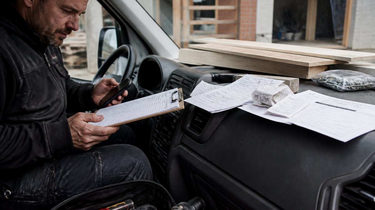 Tradesperson reviewing material invoices and job profitability data on a clipboard in a work van cab
