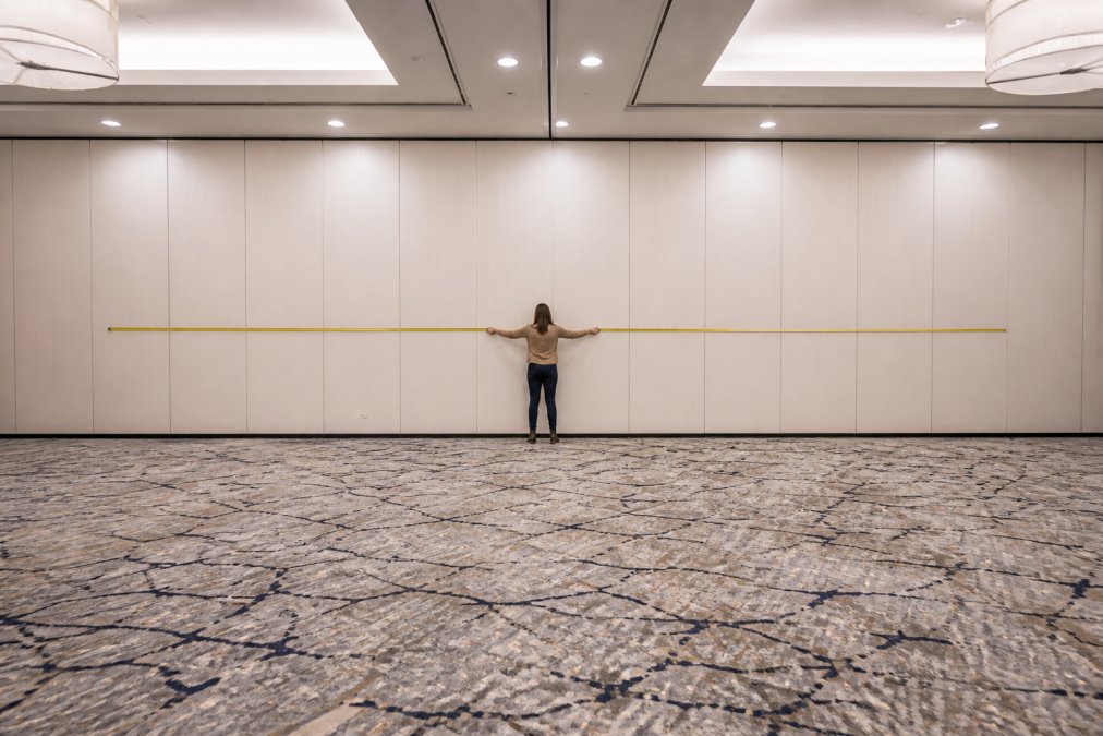 Event planner holding a yellow measuring tape stretched across the full width of an empty hotel ballroom, measuring the wall for a pipe and drape backdrop setup, bright overhead lighting, neutral walls