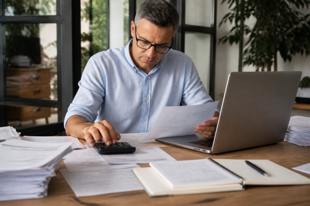 Small business owner working at desk with laptop, calculator, and financial documents including tax forms and income records, natural office lighting