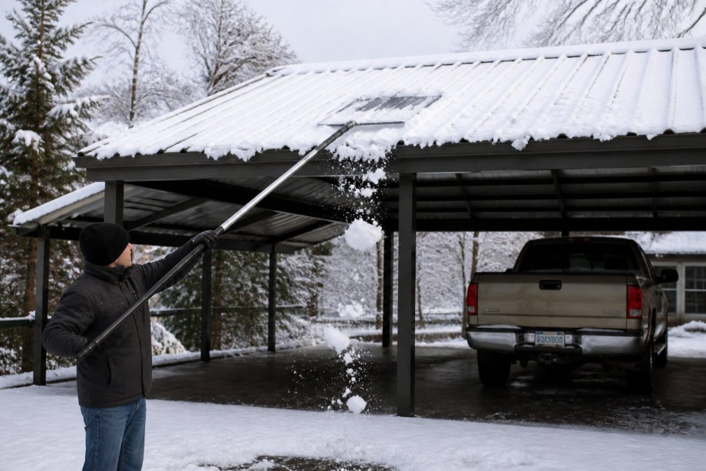 Person in winter gear using a long-handled rake to safely clear heavy snow load from a metal carport roof.