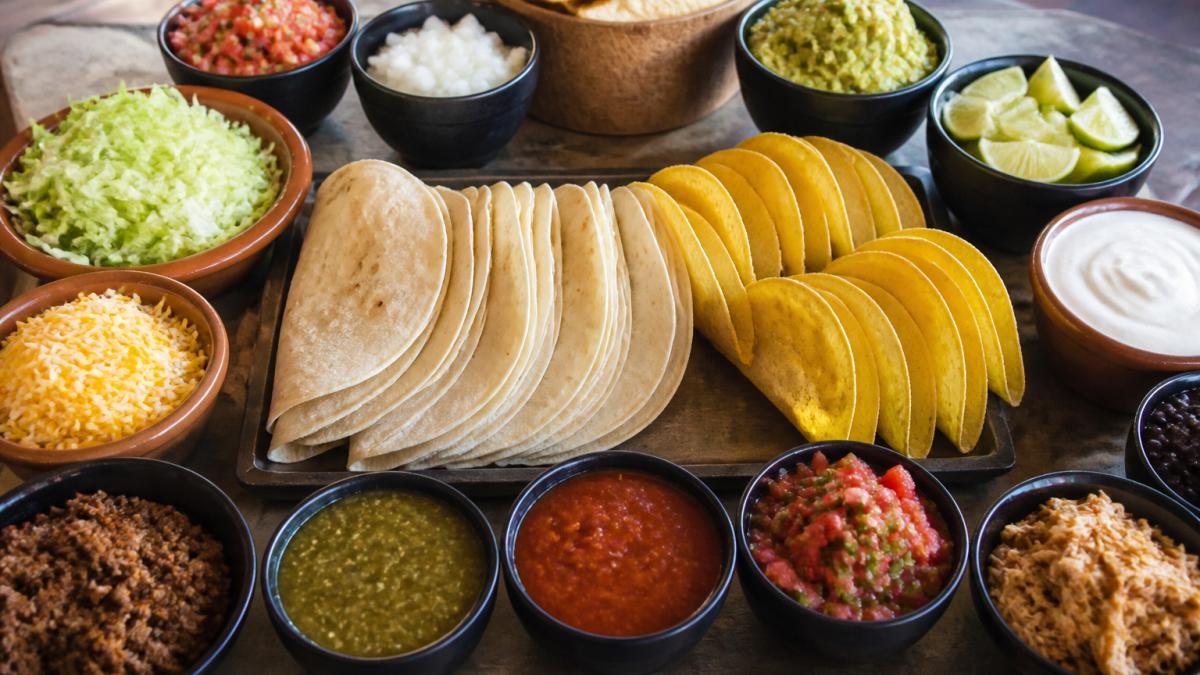 Overhead view of a fully set up taco bar with colorful bowls of shredded cheese, fresh lettuce, diced tomatoes and onions, salsa in multiple varieties, guacamole, sour cream, and lime wedges, with stacks of soft tortillas and hard taco shells arranged in the center, ready for guests to build their own tacos