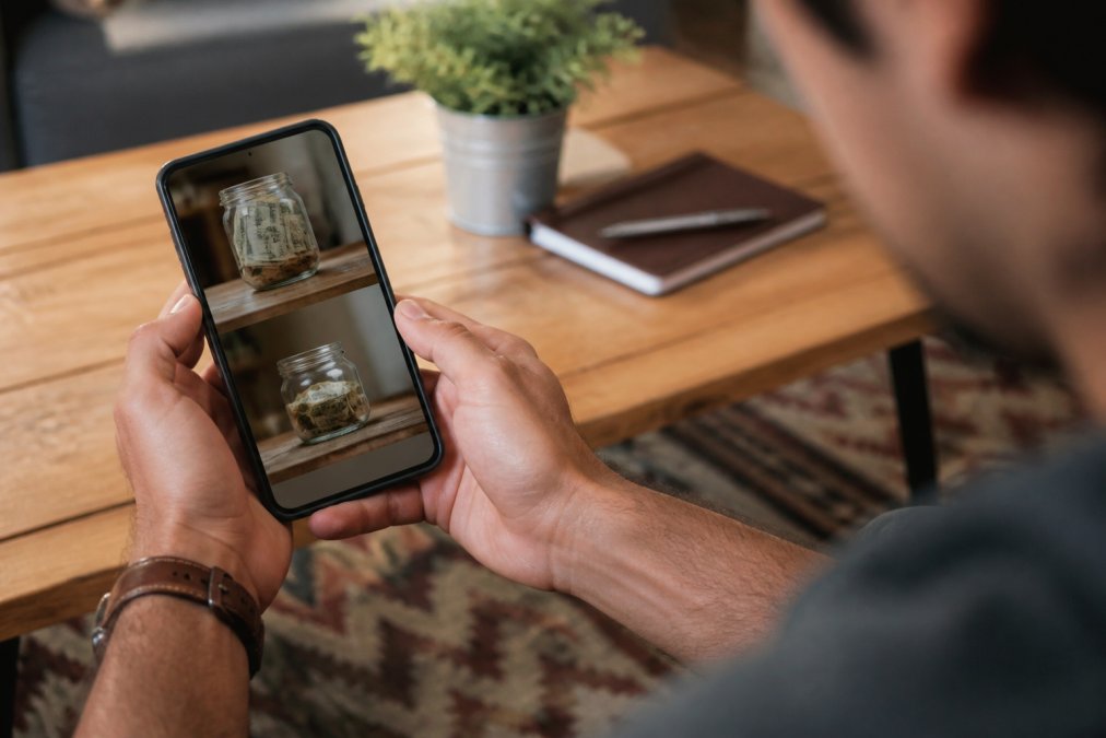 Close-up of hands holding a smartphone displaying a banking app with two savings accounts visible on screen, suggesting automated financial transfers in a clean modern setting