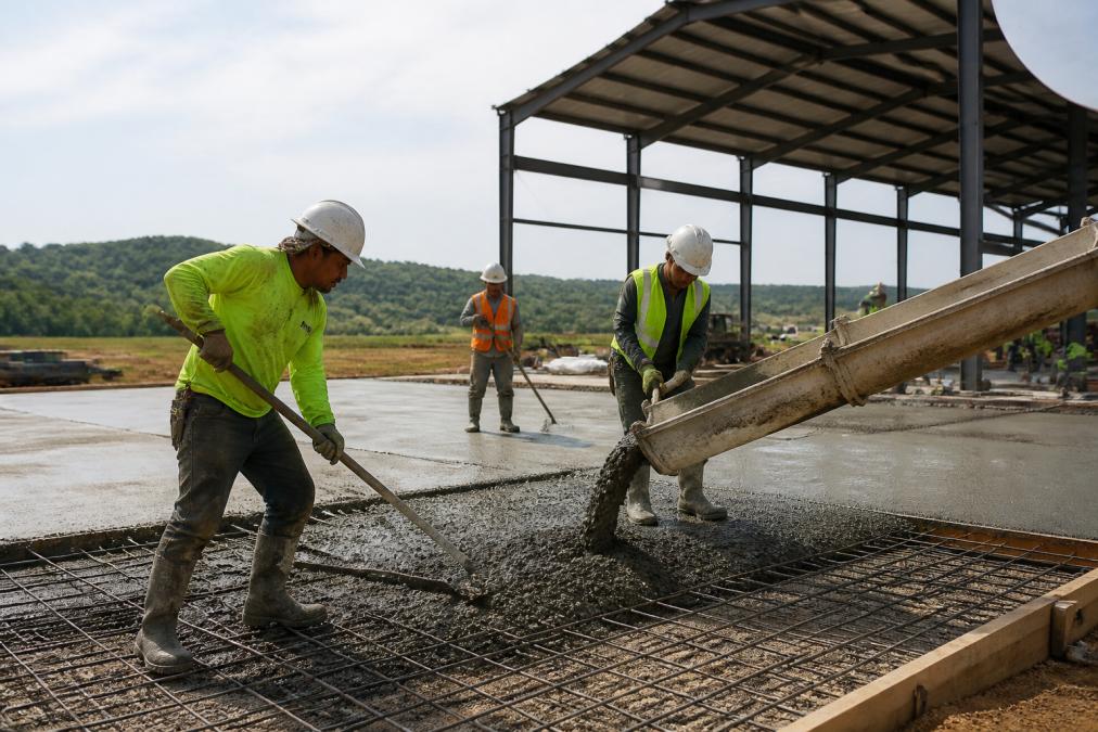 A construction crew in safety vests pouring a concrete slab foundation for a large steel building frame in a rural setting, with rebar visible and a metal building kit partially erected in the background